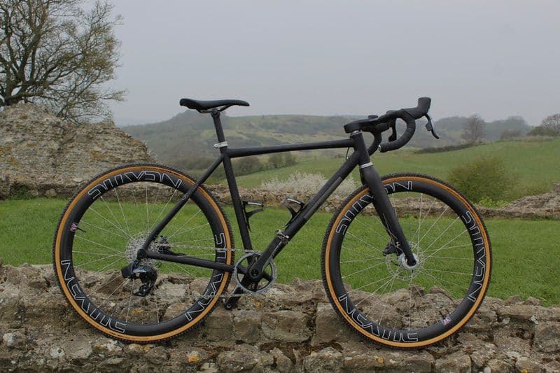 A black titanium gravel bike with tan-wall tyres in front of a stone wall
