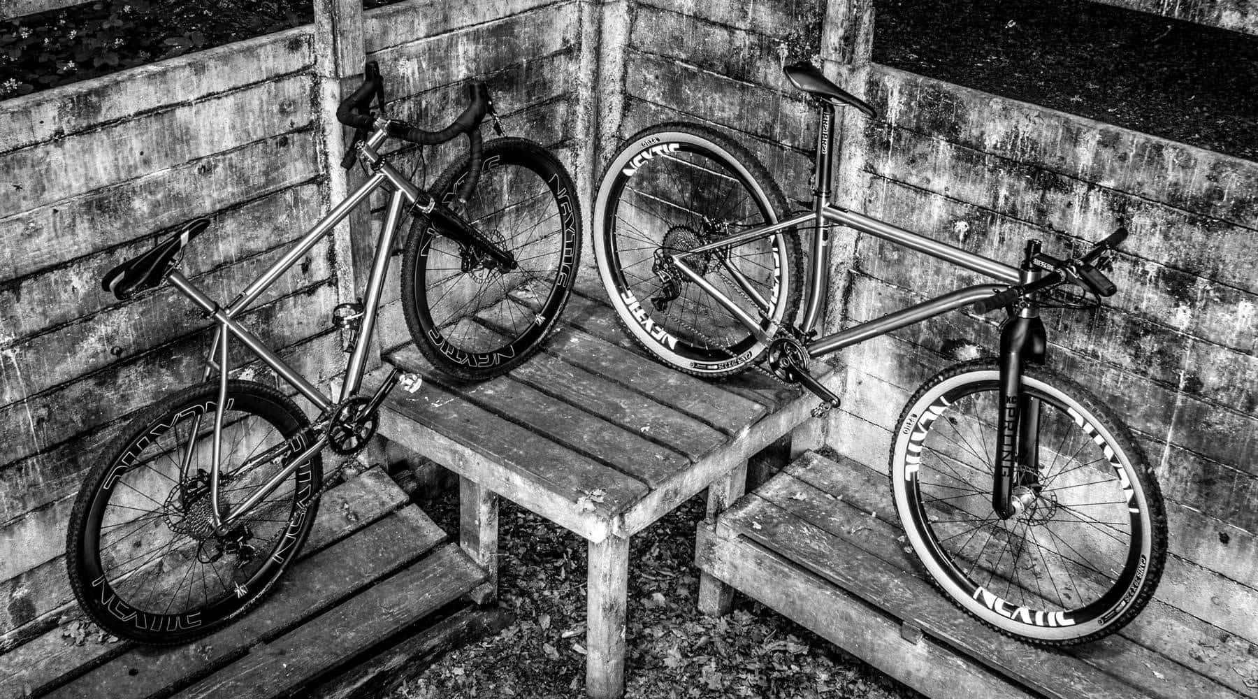 Two bikes with bare titanium frames and deep carbon wheels in a wooden shelter, in black and white